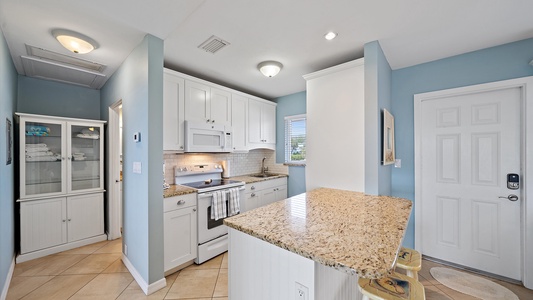 A compact kitchen with light blue walls, white cabinets, and granite countertops. There is a microwave, stove, and a small dining area with two stools. A cabinet with towels is visible in the background.