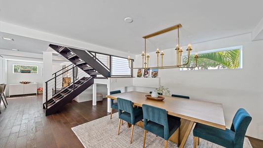 A modern dining room with a wooden table, six blue chairs, a gold chandelier, and an open staircase. The area is well-lit with natural light from the windows.