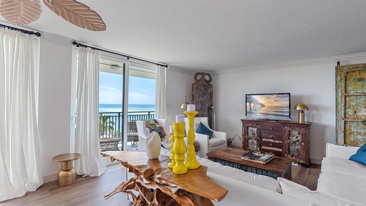 A living room with a TV on a rustic wooden stand, white sofas, a sculpted wooden coffee table, and decorative items. A sliding door opens to a balcony with ocean views. White curtains frame the door.