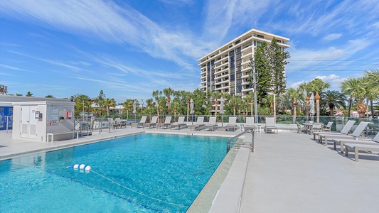 Outdoor swimming pool with lounge chairs surrounded by a concrete deck area, situated under a clear sky with a tall building and trees in the background.