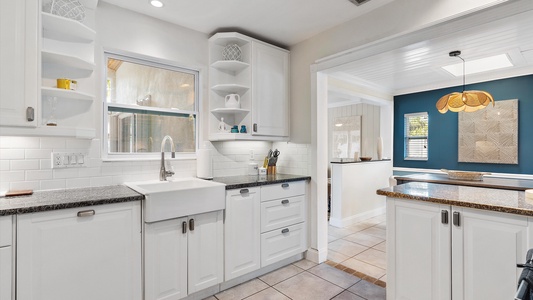 Bright kitchen with white cabinets, a farmhouse sink, stainless steel faucet, granite countertops, and open shelves. View of an adjacent room with a teal accent wall and a pendant light.