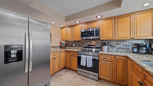 Modern kitchen with stainless steel appliances, wooden cabinets, granite countertops, and tiled backsplash. Includes refrigerator, oven, microwave, and coffee maker.