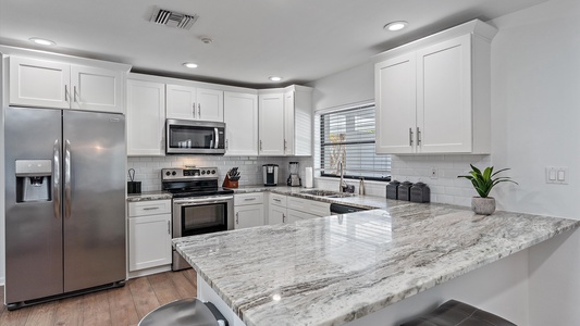 Modern kitchen with white cabinets, stainless steel appliances including a refrigerator and oven, marble countertops, a large window, and wooden flooring. A plant is placed on the counter.