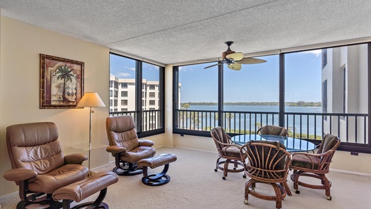 A room with large windows overlooking a body of water, featuring two leather recliners, a small round table with four chairs, a floor lamp, and a ceiling fan.