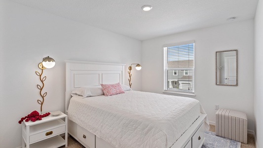 A small bedroom with a white bed frame, a white quilt, and a pink throw pillow. The room has wall-mounted lamps, a window with blinds, a wall mirror, a nightstand, and a small cushioned stool.