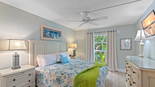 A cozy bedroom with a bed dressed in floral bedding, nightstands with lamps, a ceiling fan, a dresser, and a window with curtains overlooking greenery. There are framed pictures on the walls.