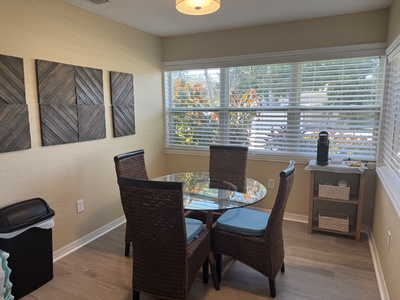 A small dining area with a round glass table and four wicker chairs. Large windows with blinds are behind the table, and there is wall art on the left. A small shelf with items is on the right.