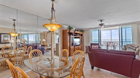 A spacious, well-lit living room with a glass dining table, wooden chairs, two sofas, a wooden TV cabinet, and large windows offering an ocean view. A mirrored wall enhances the room's brightness.