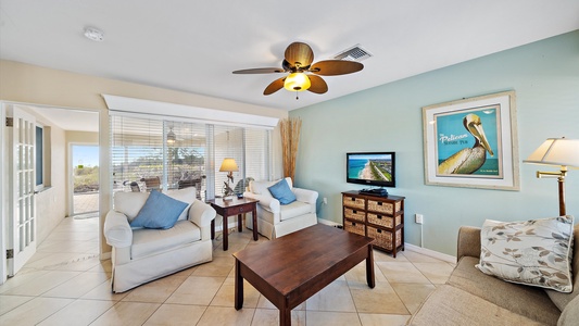 A cozy living room with a ceiling fan, two white armchairs, a beige sofa, a wooden coffee table, and a TV on a wicker stand. There's a large window and a door leading to a well-lit hallway.