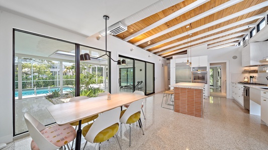 Bright modern kitchen and dining area featuring a wooden ceiling with exposed beams, large windows, and sliding glass doors leading to an outdoor pool.