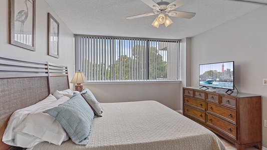 A bedroom with a large bed, wooden headboard, two framed pictures, a window with vertical blinds, a ceiling fan, a dresser with a TV, and a table lamp.