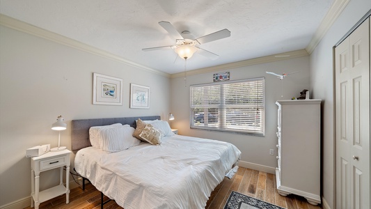 A small bedroom with a bed, two nightstands, a dresser, and a ceiling fan. The room has light-colored walls, wood flooring, a window with blinds, and framed artwork on the walls.