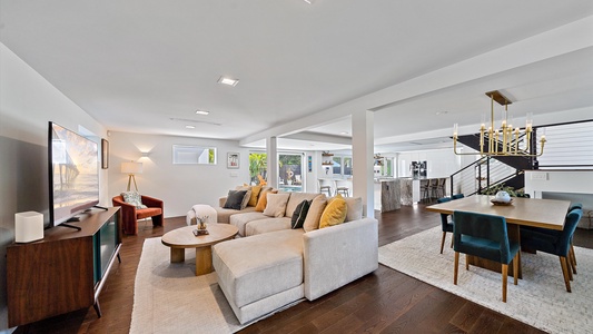 A modern open-concept living space features a beige sectional sofa, a wooden coffee table, a flat-screen TV, a dining table with green chairs, and a kitchen in the background with a breakfast bar.