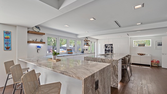 Modern kitchen interior with large marble countertops, white cabinets, an island with built-in stovetop, bar stools, and ample natural light from large windows.