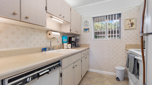 A small kitchen with beige cabinets, a dishwasher, a sink, a countertop with a coffee maker, wall clock, and window with blinds. A stove and a trash bin are also visible.