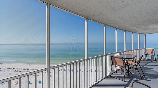 A curved balcony overlooking a serene beach with blue water. The balcony has a table and chairs, and is enclosed with floor-to-ceiling glass panels.