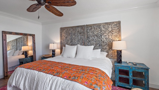 A cozy bedroom with a large bed adorned with white pillows and a colorful throw blanket, two blue nightstands with lamps, and a large decorative mirror against the wall. Ceiling fan is visible.