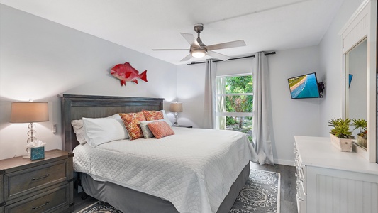 A bedroom featuring a large bed with white bedding, two nightstands with lamps, a mounted fish decoration, a ceiling fan, a wall-mounted TV, a dresser with decor, and a window with white curtains.