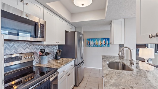 A modern kitchen with stainless steel appliances, white cabinetry, a marble countertop, and a chevron tile backsplash. A sailboat painting decorates the wall near the refrigerator.