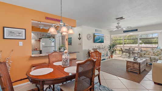 A living and dining area with a round wooden table and four chairs, a kitchen with white appliances, a seating area with a couch and TV, and a glass sliding door leading to an outdoor view.