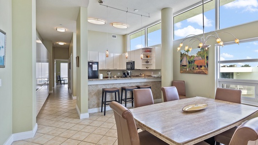 A modern, well-lit kitchen and dining area with high ceilings, tile flooring, and large windows. The space includes a dining table with chairs, bar stools at the kitchen counter, and light fixtures.