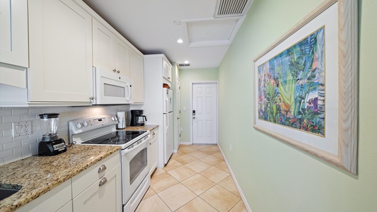 A narrow kitchen with white cabinets, granite countertops, and stainless steel appliances. A vibrant painting hangs on the pale green wall. The floor is tiled, and a door is visible at the end.