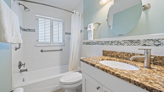 A modern bathroom with a granite countertop, single sink vanity, white bathtub with curtain, white tiled walls, and a window with shutters. There is a towel bar, overhead light, and round mirror.