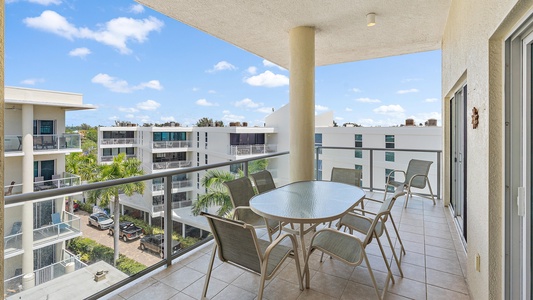 A spacious balcony with a glass-top table and six chairs overlooks modern apartment buildings and trees under a blue sky with scattered clouds.