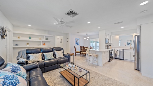 A bright, open living room with a black leather sofa, glass coffee table, and built-in shelves. Adjacent is a white kitchen with an island and bar stools. A dining area with a round table is in the background.