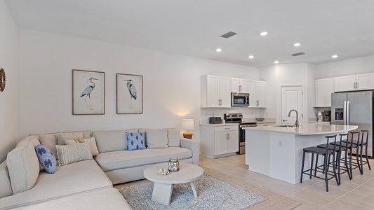 Modern open-concept living area with a white L-shaped sofa, coffee table, two bird-themed wall artworks, a kitchen with white cabinets, stainless steel appliances, and a kitchen island with barstools.