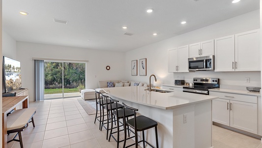 A modern kitchen with white cabinetry and a large island featuring black stools. The space opens into a living area with a couch and a TV, with large glass doors leading to a patio and greenery outside.