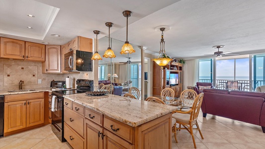 A modern open-plan kitchen and living area with a granite-topped island, wooden cabinets, a dining table, and a sitting area. Large windows provide a view of the ocean from the balcony.