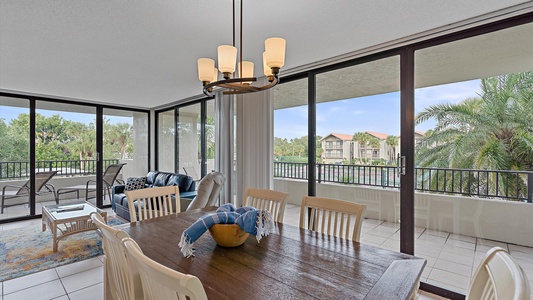 A dining area with a wooden table and white chairs opens to a living space with a sofa. Large windows and sliding doors reveal an outdoor balcony with seating and views of trees and buildings.