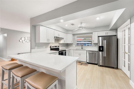 Modern kitchen with white cabinetry, stainless steel appliances, and a breakfast bar with three beige stools. Light wood flooring and a ceiling fan are also visible.