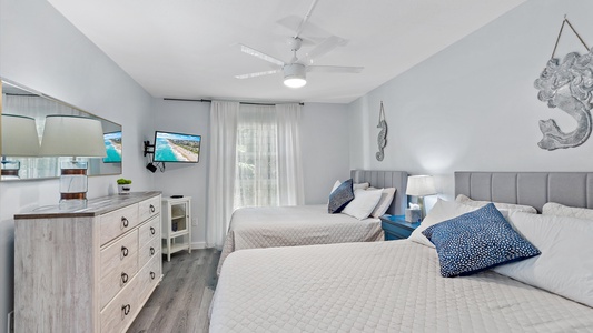 A bedroom with two double beds, a dresser with a mirror, a TV, and a ceiling fan. The room is decorated with blue accents, seashell wall art, and has a large window with white curtains.