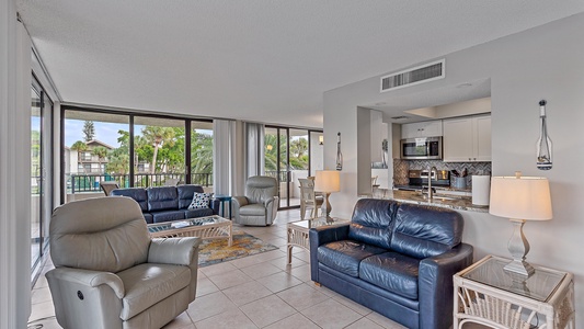 Modern living room with tiled floor, featuring two leather sofas, two recliner chairs, and a glass coffee table. The room has large windows, a view of a balcony, and an adjacent open kitchen area.