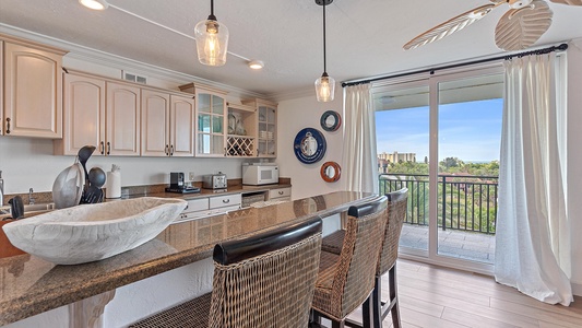 A well-lit kitchen with beige cabinets, a dark granite counter with two wicker bar stools, nautical decor, and glass doors opening to a balcony with a view of greenery and buildings.