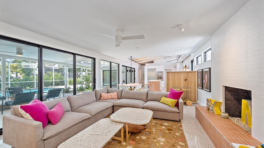 A modern living room with a large L-shaped sofa, colorful cushions, coffee tables, and a brick fireplace. Large windows offer a view of an outdoor pool area.