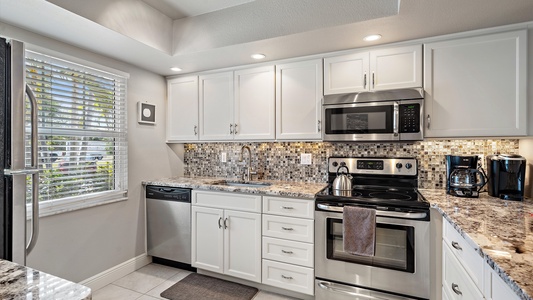 A modern kitchen with stainless steel appliances, white cabinets, a speckled granite countertop, mosaic tile backsplash, a window with blinds, and under-cabinet lighting. A towel hangs on the oven.