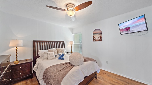 A bedroom with a ceiling fan, a wooden bed with pillows and a blanket, nightstand with a lamp, a dresser, wall art, a window, and a wall-mounted television.