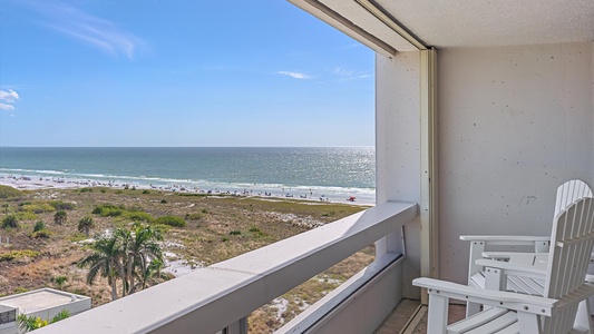 View of a beach and ocean from a balcony with white Adirondack chairs, overlooking greenery, sandy shore, and scattered beachgoers under clear blue skies.