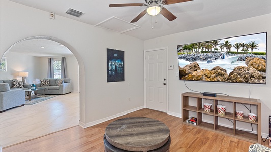 A living room area with a wall-mounted TV displaying a tropical scene, a ceiling fan above, and an archway leading to a sitting area with sofas in the background. There is also a round coffee table.