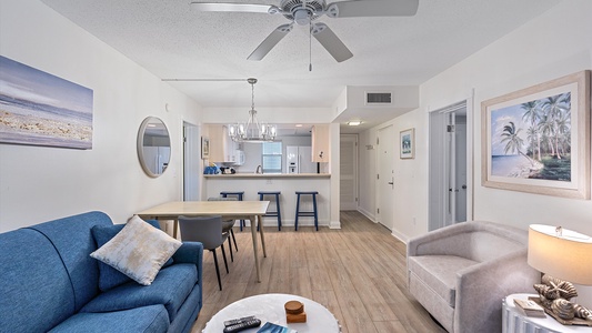 A neatly arranged living room with a blue sofa, a wooden dining table, bar stools at a kitchen counter, and beach-themed artwork. The room is brightly lit with a ceiling fan overhead.