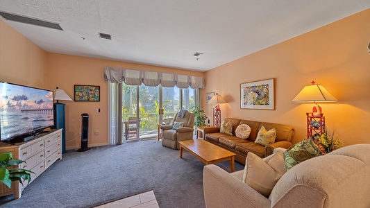 A cozy living room with peach walls, a large TV, a sofa, armchairs, a coffee table, and lamps. Large windows with curtains provide natural light and a view of the greenery outside.