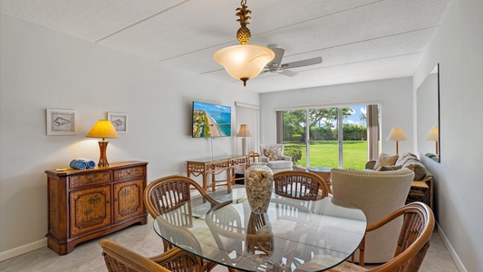 A living room features a glass dining table with wicker chairs, a TV mounted on the wall, a cushioned armchair, a wooden sideboard, table lamps, and a large window overlooking a grassy outdoor area.