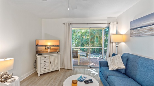 A small living room with a blue sofa, a TV on a white stand displaying a sunset image, a round coffee table, and a sliding glass door opening to a patio with greenery outside.