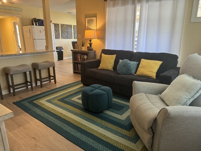 A cozy living room with a dark sofa, light armchair, patterned rug, and two bar stools by the kitchen counter. Decor includes pillows, a lamp, and wall art. Light filters through white curtains.