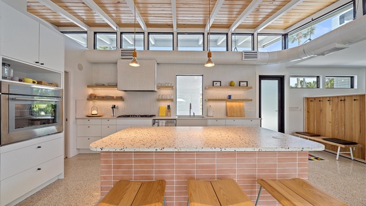 A modern kitchen with a terrazzo island, wooden stools, white cabinetry, built-in appliances, open shelving, and large windows. A row of wooden lockers is visible on the right.