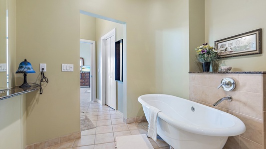 A bathroom with a freestanding bathtub, light-colored walls, tiled floor, and a marble counter with a blue lamp and a wall-mounted hair dryer. A doorway leads to another room.