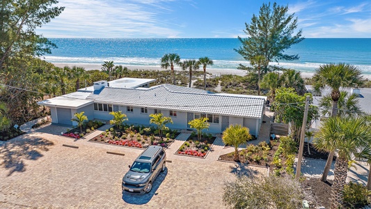 A single-story beachfront house with a gray roof and a landscaped front yard, featuring a single SUV parked in the driveway. The ocean is visible in the background under a blue sky.